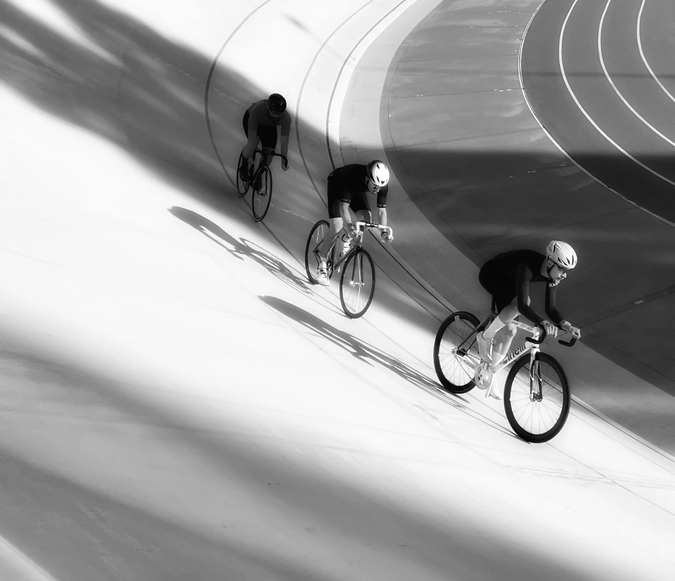 Three cyclists on the banked curve of a velodrome, black and white