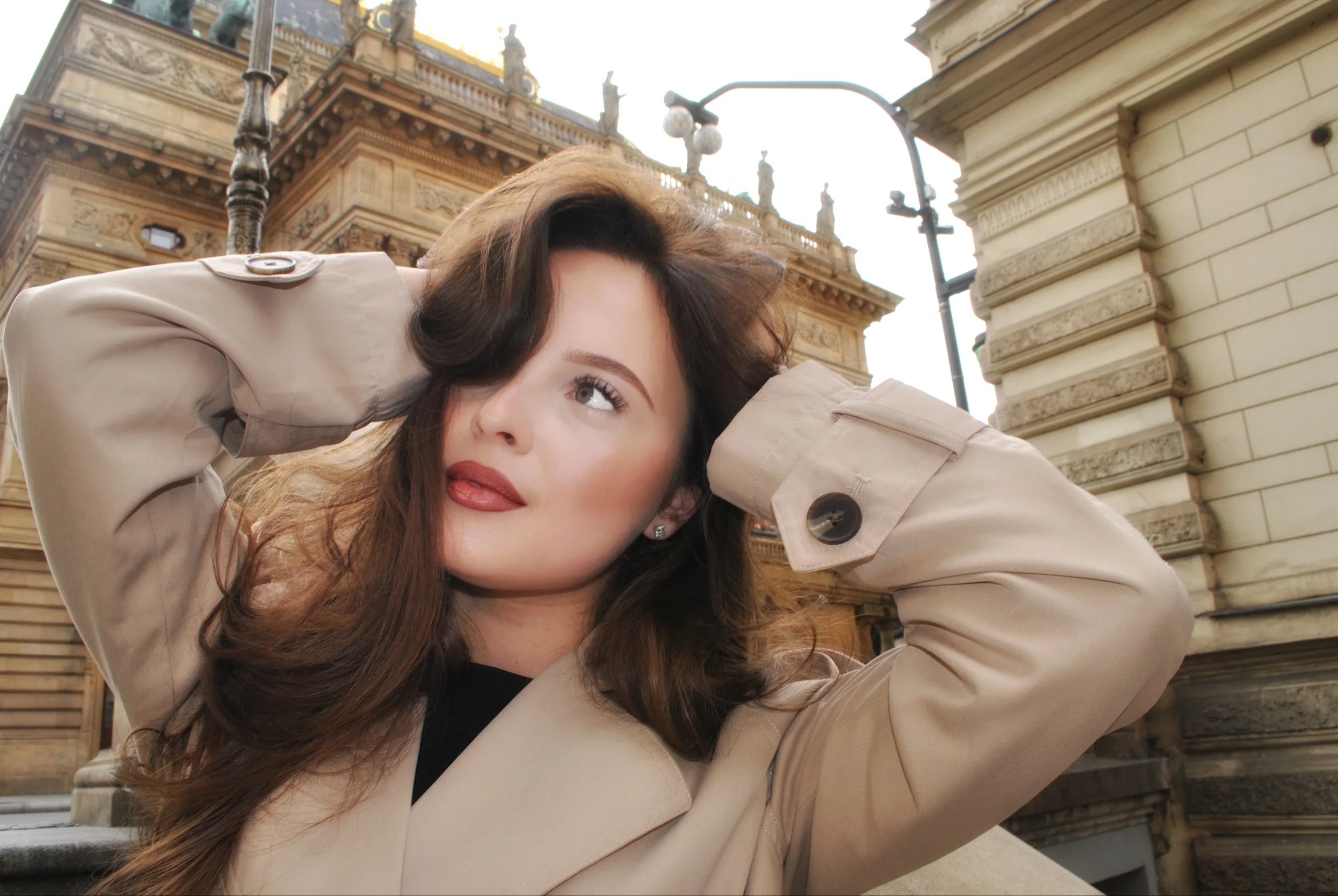 Women's portrait: beige trench coat, hands in her hair, historic buildings of Prague