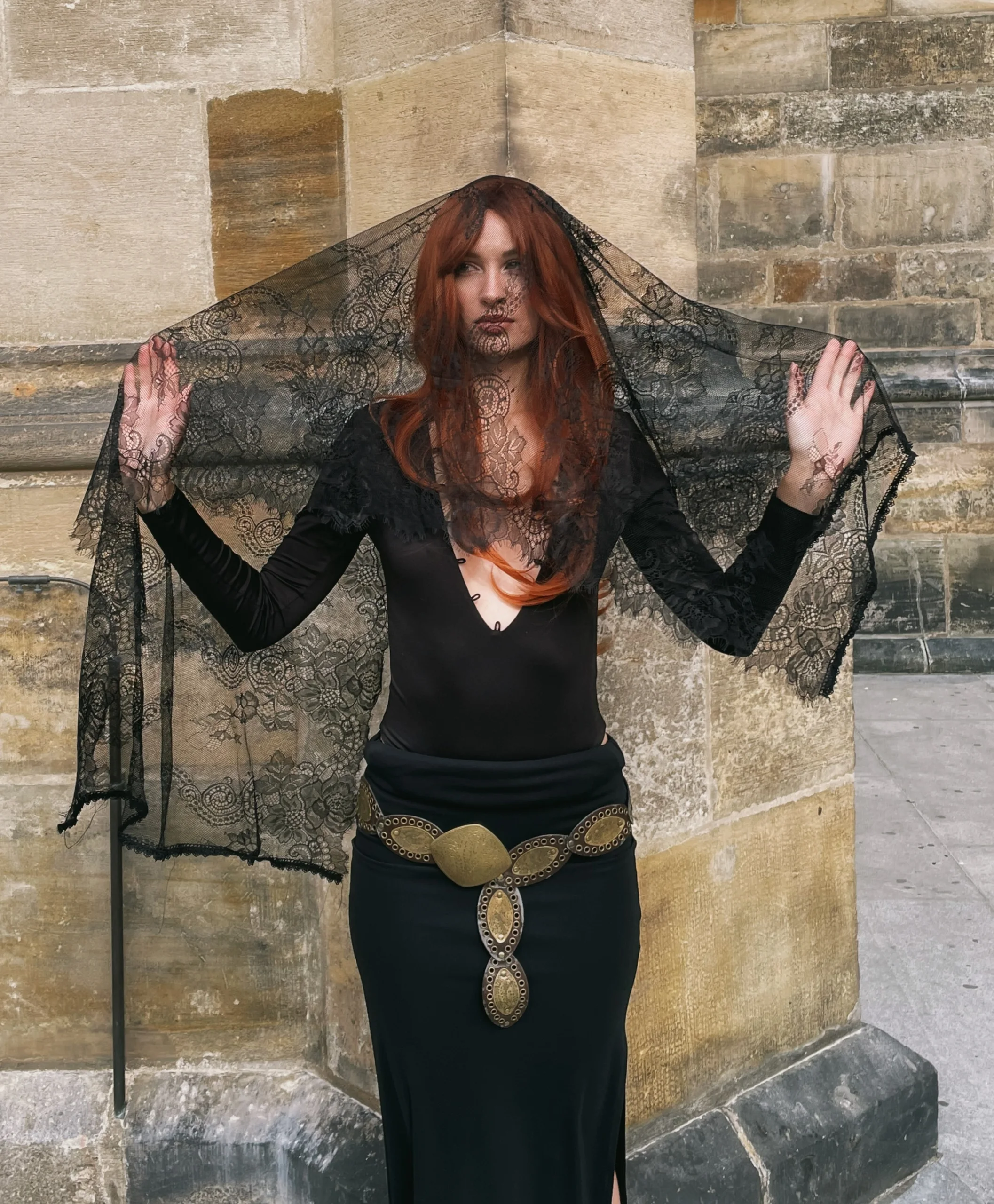 Red-haired model in black holding a lace veil over her head against a Gothic stone wall